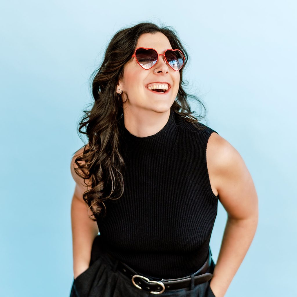 Caitlin dressed in all black, wearing red heart shaped glasses, laughing on a blue background.